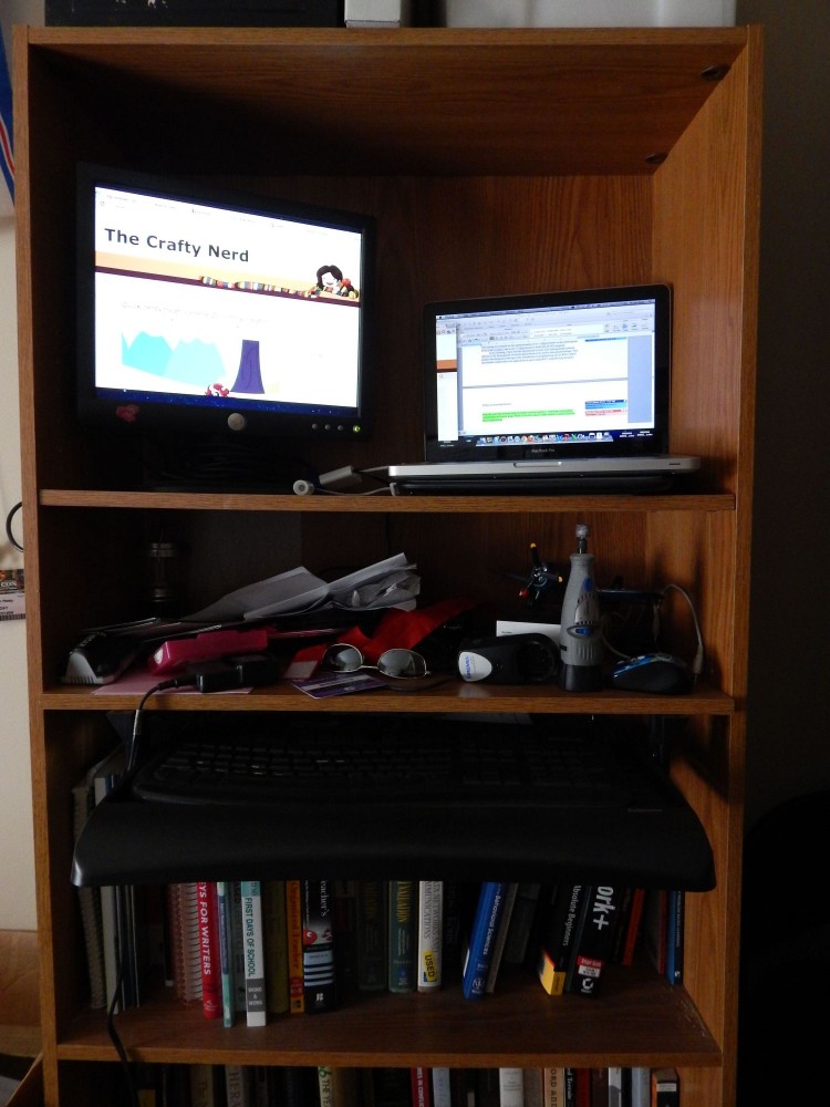 Image of a bookshelf with a keyboard drawer attached to one of the shelves, and a laptop with external monitor on the top shelf.