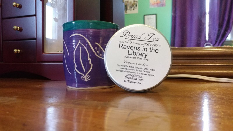 Photo of a handmade mug, with a tin of tea labeled Ravens in the Library.
