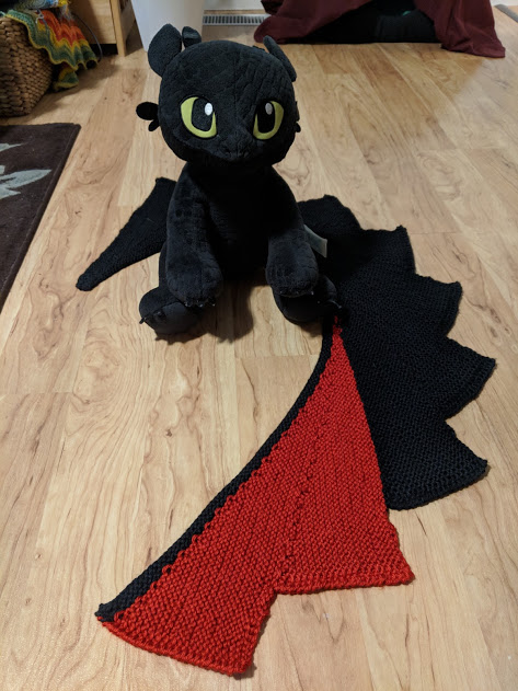 Plush Toothless the Dragon, sitting on the floor next to a shawl that resembles a dragon's wing.