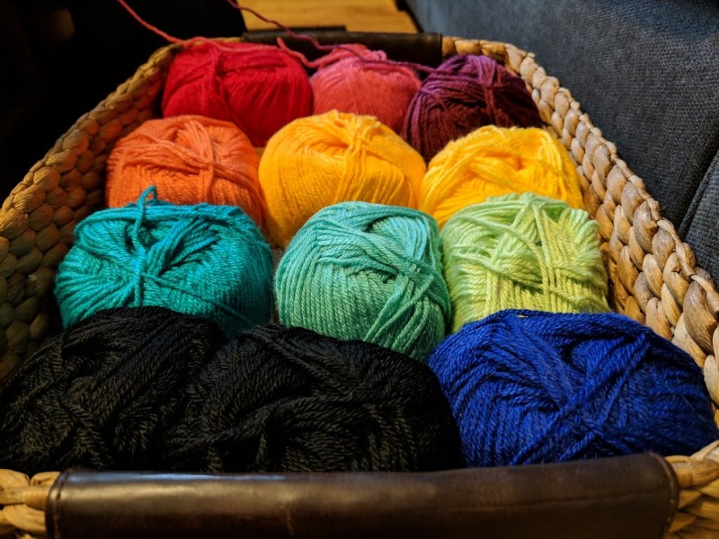 Basket of 12 skeins of yarn - 10 of the skeins are in various rainbow hues, while the remaining two are black.