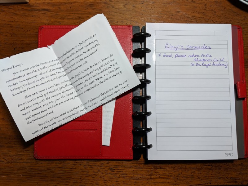 Red notebook with removable pages. There's a letter to Ëlinyr laying on the left side of the notebook.