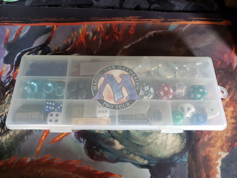 Large plastic container with the Magic: The Gathering logo on the front, holding lots of dice, health counters, and glass pebbles that are used as counters.