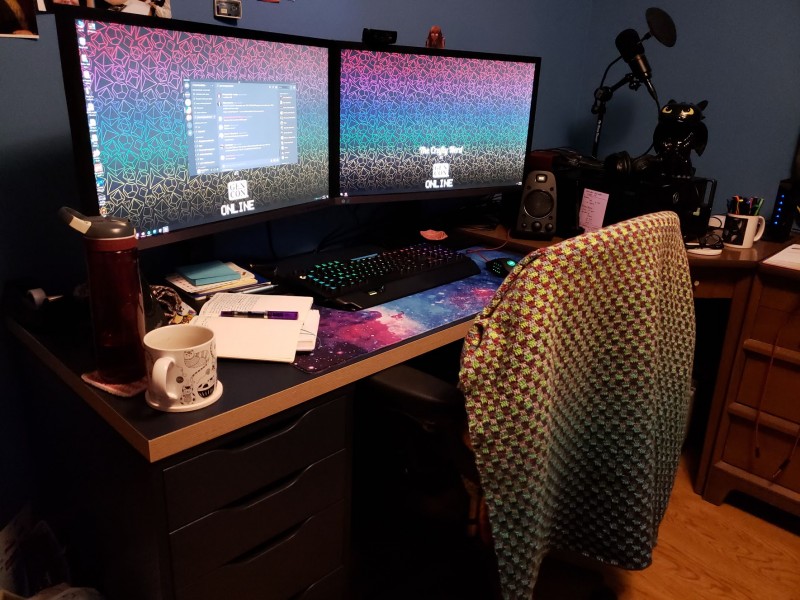 A desk with two large monitors and a desktop computer on it. The computer screens have a custom Crafty Nerd at Gen Con wallpaper.