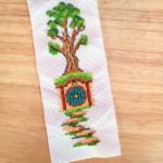 Photograph of a cross-stitch bookmark featuring a door to a hobbit house underneath a hill with a tree growing on it.