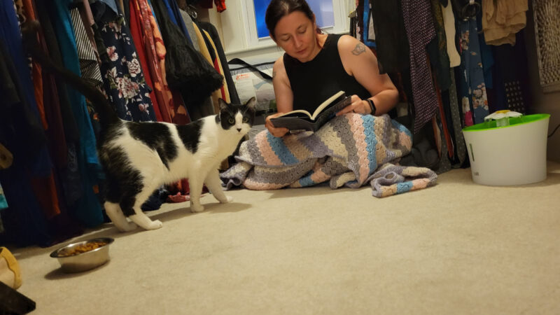 Photograph of me reading the book "The Slow Regard of Silent Things" to Anya, a small black and white cat. Anya and I are sitting in a large walk-in closet. 