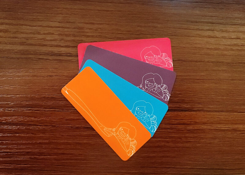 Picture of four mini business cards, half the height of a typical business card, in orange, blue, purple, and pink. Each business card has white line art of the old Crafty Nerd logo, and it's hard to see over the bright colors.