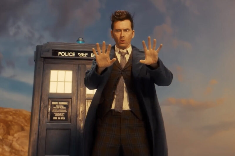 David Tennant as the Fourteenth Doctor standing in front of the TARDIS, looking at his hands with an almost confused expression on his face.